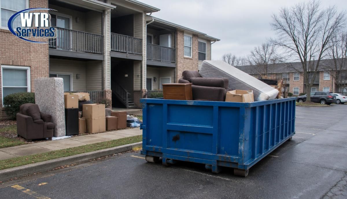 Blue WTR Services roll-off dumpster outside a multi-unit apartment building during tenant move-out, loaded with furniture, mattresses, and boxes